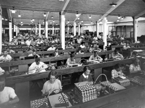 Interiörbild från verkstad där kvinnor och män och manliga sitter i rader vid långa arbetsbord.