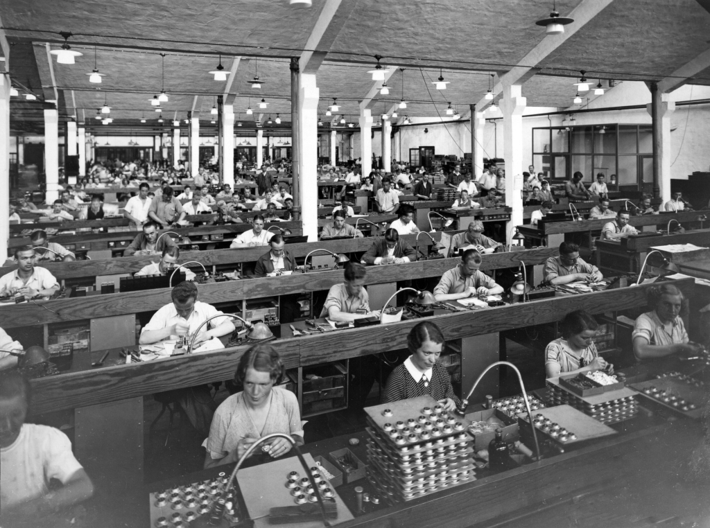 Interiörbild från verkstad där kvinnor och män och manliga sitter i rader vid långa arbetsbord.