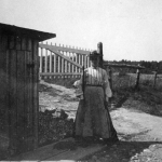 Svartvitt fotografi, kvinna står bredvid liten bod, i bakgrunden grind vid järnvägsövergång.