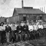 Gruppfotografi, drygt 20 arbetsklädda män står uppställda i två rader framför trähus.