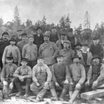 svartvitt foto av tjugotvå män uppställda framför brädhögar.