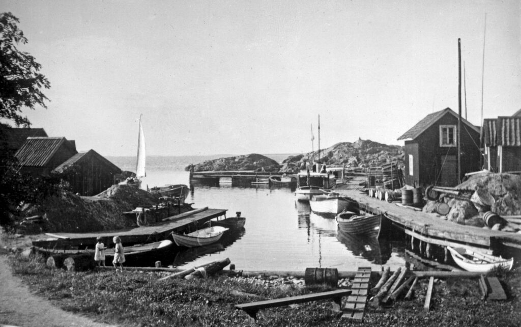 Svartvit bild av fiskeläget med små bodar, träbåtar och bryggor. Två barn leker vid vattnet.