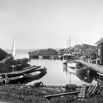 Svartvit bild av fiskeläget med små bodar, träbåtar och bryggor. Två barn leker vid vattnet.