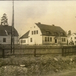 Svartvit bild med fyra vita byggnader. En stor i mitten omgiven av mindre hus.