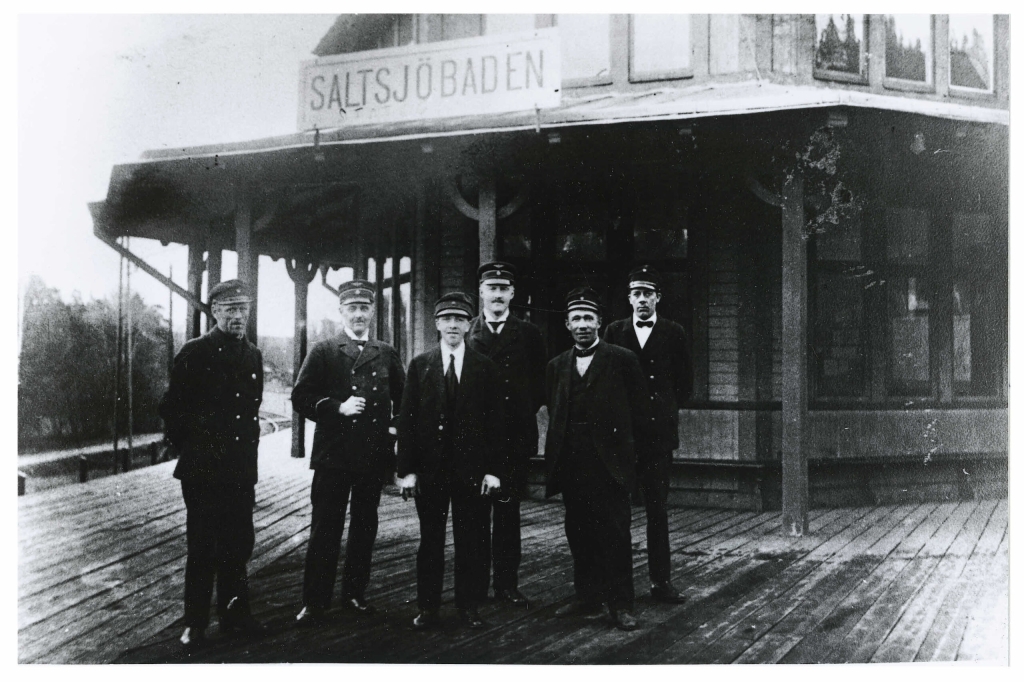 Svartvitt fotografi. 6 män i arbetskläder, uniform, står uppställda framför Saltsjöbadens gamla stationshus.