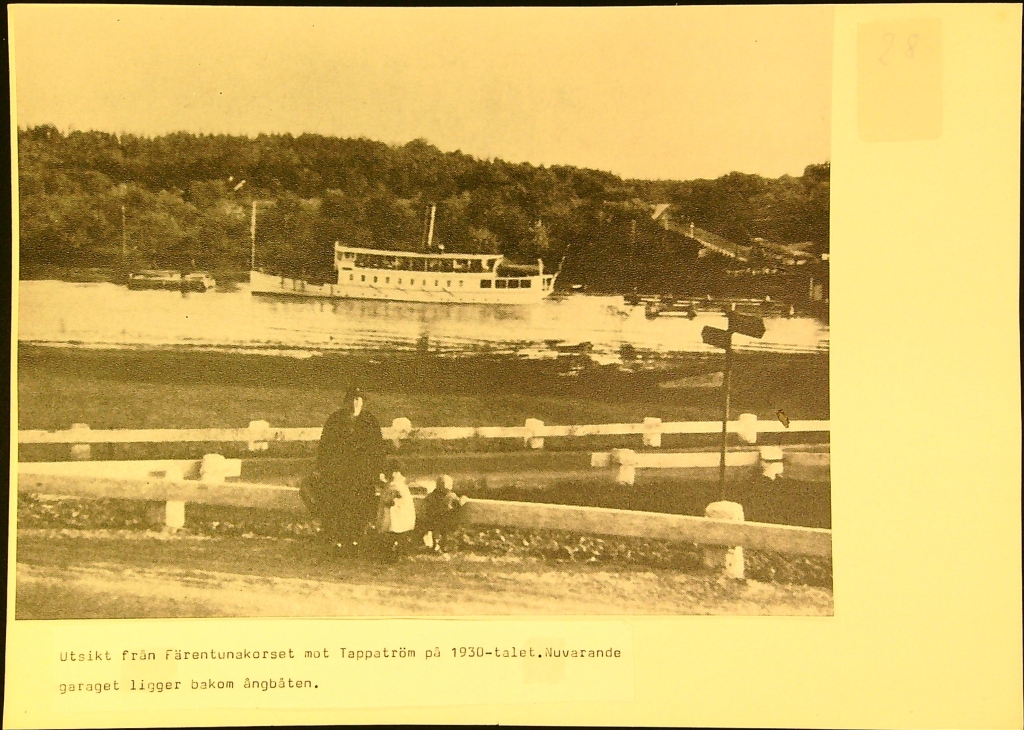 En kvinna och två barn står mitt i bilden. Bakom den syns en ångbåt på vattnet och på andra sidan vattnet skog och lite av en bro.