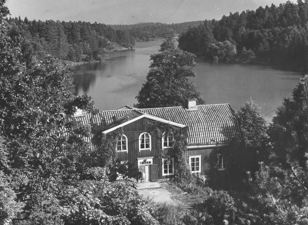 Svartvitt fotografi, stort trähus, i bakgrunden vy över vatten.