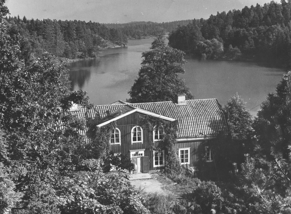 Svartvitt fotografi, stort trähus, i bakgrunden vy över vatten.
