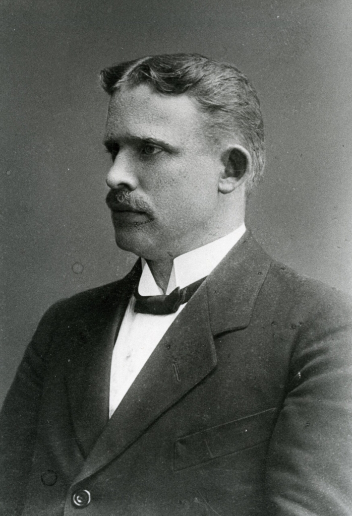 Svartvitt fotografi av en man i profil som bär fluga och mustasch.