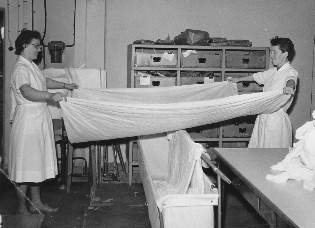 Svartvitt fotografi, två kvinnor står och sträcker och viker lakan i tvätteriet.