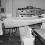 Svartvitt fotografi, två kvinnor står och sträcker och viker lakan i tvätteriet.