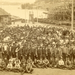 Ett hundratal män står uppställda på skeppsvarvets. Ett tjugotal pojkar sitter i skräddarställning längst fram. I bakgrunden syns byggnadsställningar av trä och havet.