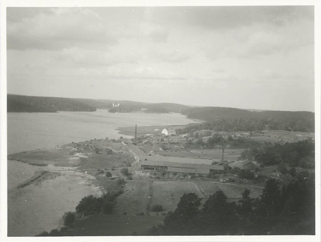 Vy från hög höjd över vatten till vänster och land till höger där några hus syns i mitten, bland dem långa lador och ett vitt hus på en höjd. I bakgrunden skymtar en vit byggnad längst in i viken.