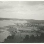 Vy från hög höjd över vatten till vänster och land till höger där några hus syns i mitten, bland dem långa lador och ett vitt hus på en höjd. I bakgrunden skymtar en vit byggnad längst in i viken.