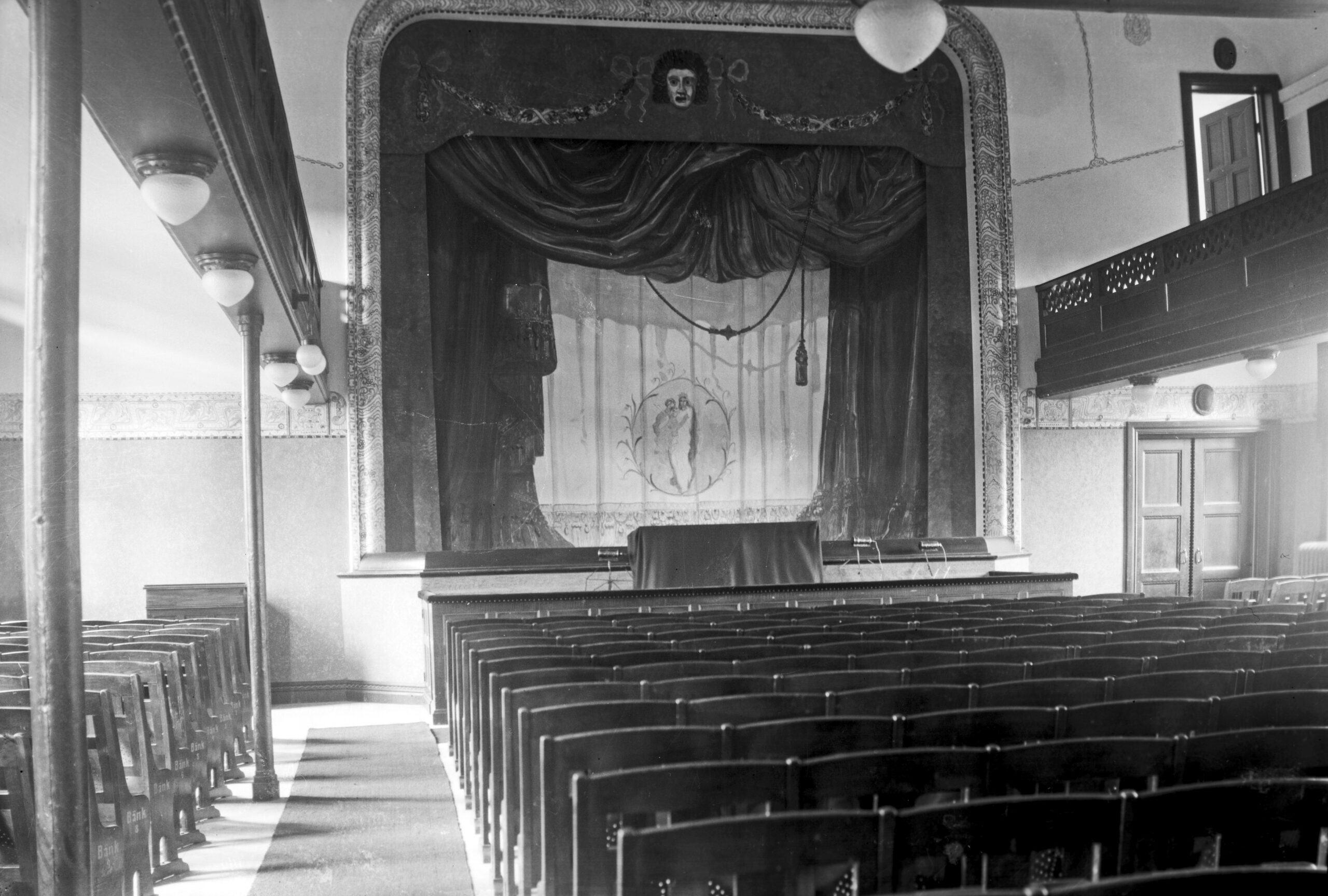 En scen med målade kulisser, dekorer och ridå. Golvet är platt och har många stolsrader i tre sektioner. Lokalens långsidor har balkonger.