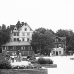 Barn och kvinnor på ett torg med planteringar och bänkar. I bakgrundens syns tre trähus, ett ligger på en kulle och har fyra våningar och ett torn. De andra ligger mot gatan i torgets fond. Det ena huset har fyra garageportar mot vägen och det andra har en skylt där det står "skor" och "läder".