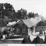 En svartvit bild av det lilla stationshuset där ett rykande ånglok åker på rälsen bortom huset. Bilden är ett vykort och har en text skriven i nederkanten "Nynäs hafsbad. Järnvägsstationen."