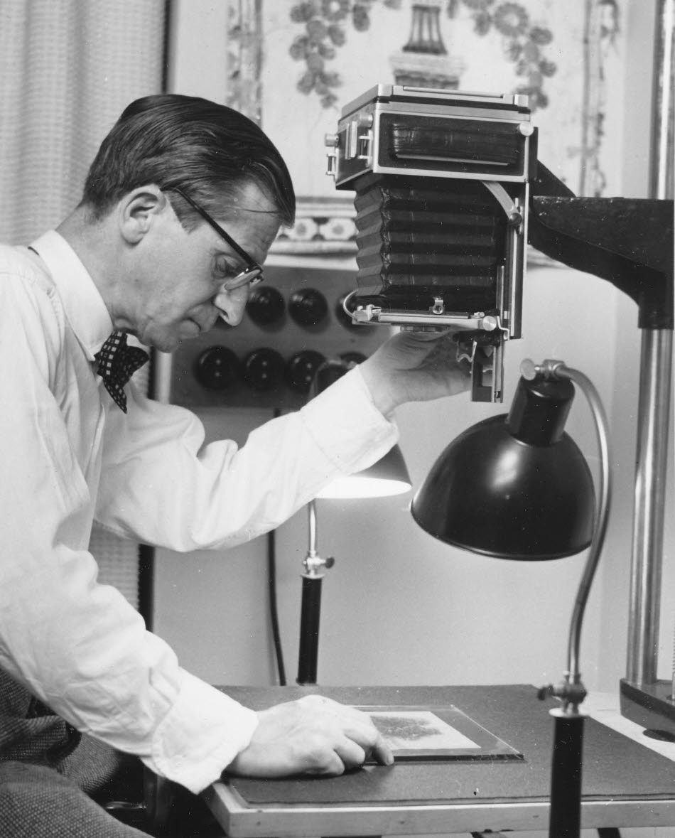 Ett svartvit foto av Ericsson med vit skjorta, fluga, glasögon och välfriserat hår som riktar kamerautrustning mot en bild på bordet. Två lampor lyser på motivet men vinkeln gör det svårt att urskilja.