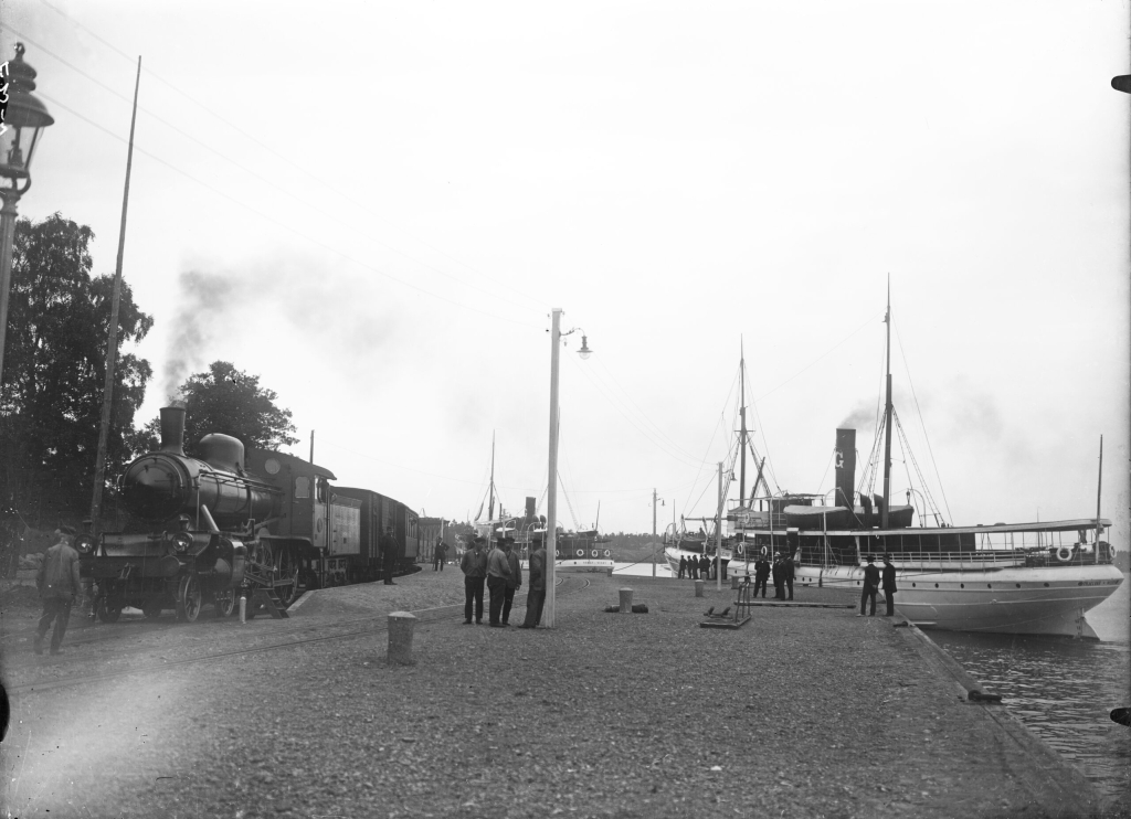 Ett ånglok har stannat vid perrongen på kajen. Två båtar ligger vid kajen. Det ryker ur både lokets och ångbåtarnas skorstenar.