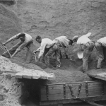 Fem män står böjda över en sandgrop och gräver upp sand.