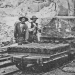 foto av två män i hatt och mustasch vid en vagn på räls. På vagnen står ett stenblock.