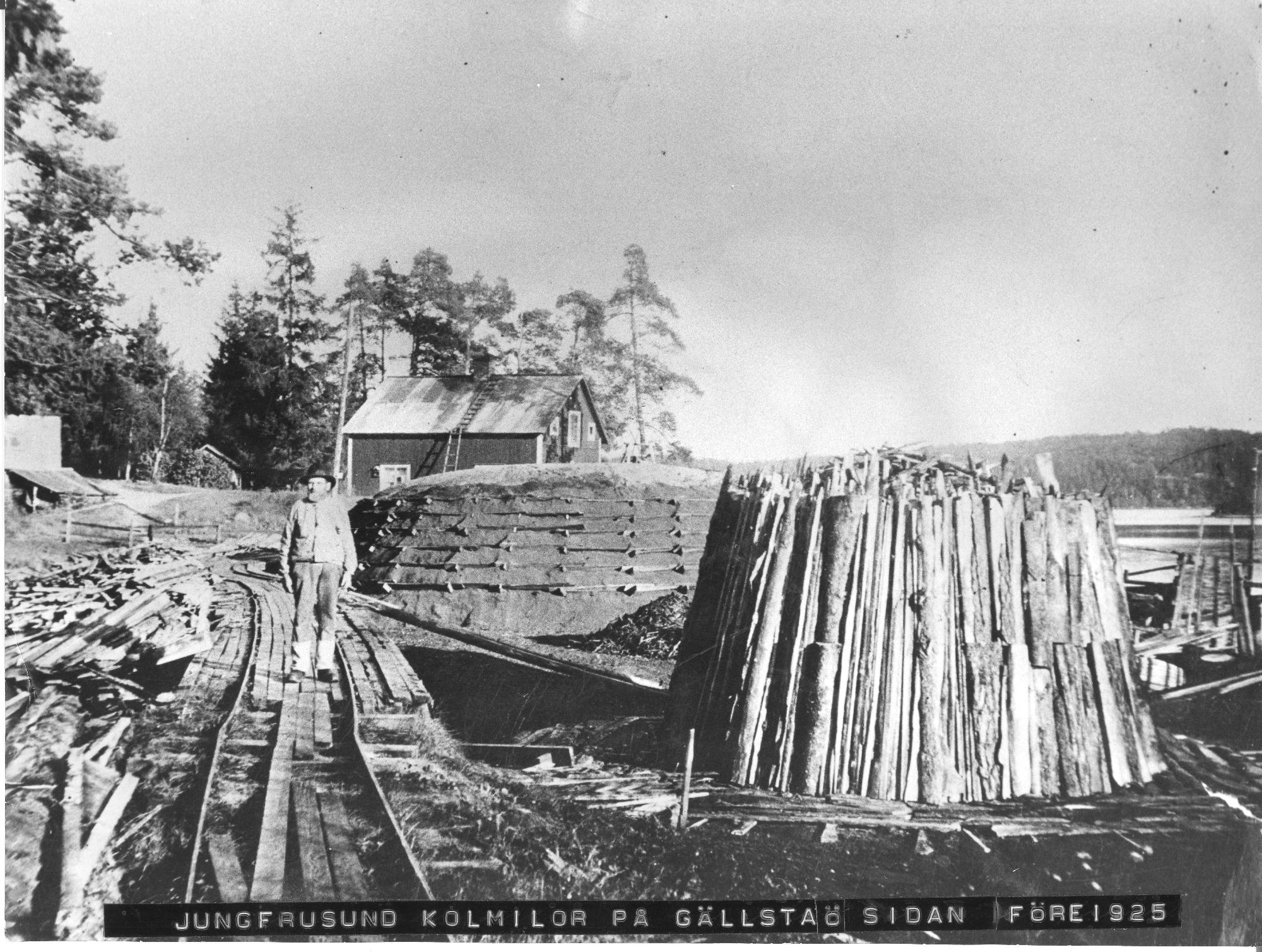 Svartvitt foto av en man med hatt som står mitt på en räls som går förbi två kolmilor intill vattnet.