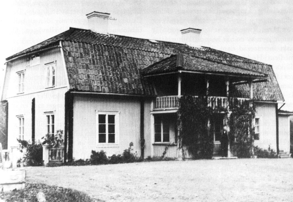 Ett svartvitt foto av en mangårdsbyggnad i trä med två våningar, brutet tak och en veranda med klätterväxter. En man står i entrén.