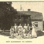 Tretton flickor står uppställda framför Glömsta gård tillsammans med sin föreståndarinna. Texten under bilden lyder: Från barnhemmet vid Glömsta.