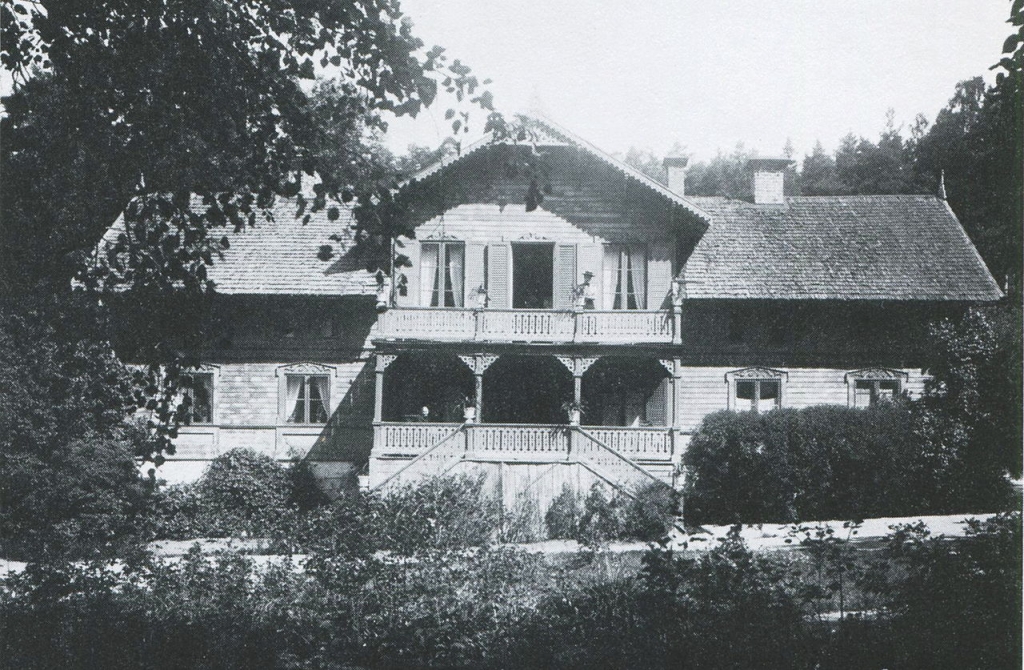 Ett trähus med snickarglädje. En kvinna i hatt står på balkongen.