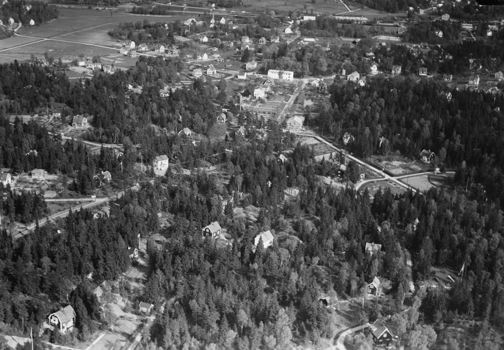 Ett svartvitt flygfoto med villor som sticker fram i skogen. Längre bort syns öppnafält och järnväg.