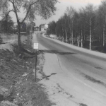 Ett svartvitt foto av en landsväg som leder upp för en backe till några hus. Mitt i bilden står en skylt.