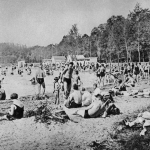 Svartivtt foto av ett hundratal människor på en sandstrand.