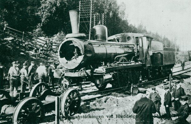 En svartvit bild av människor på och runt spåret där ett trasigt ånglok står.