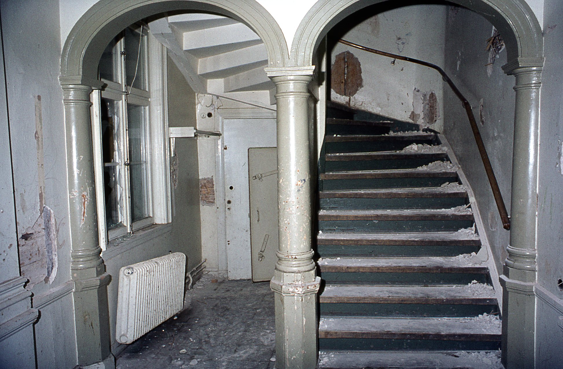 Ett trapphus målat i vitt med högar av murbruk på trappans steg. Ett element hänger på sned, färgen är avskavd på de svarvade träpelarna och väggarna är fulla av hål.
