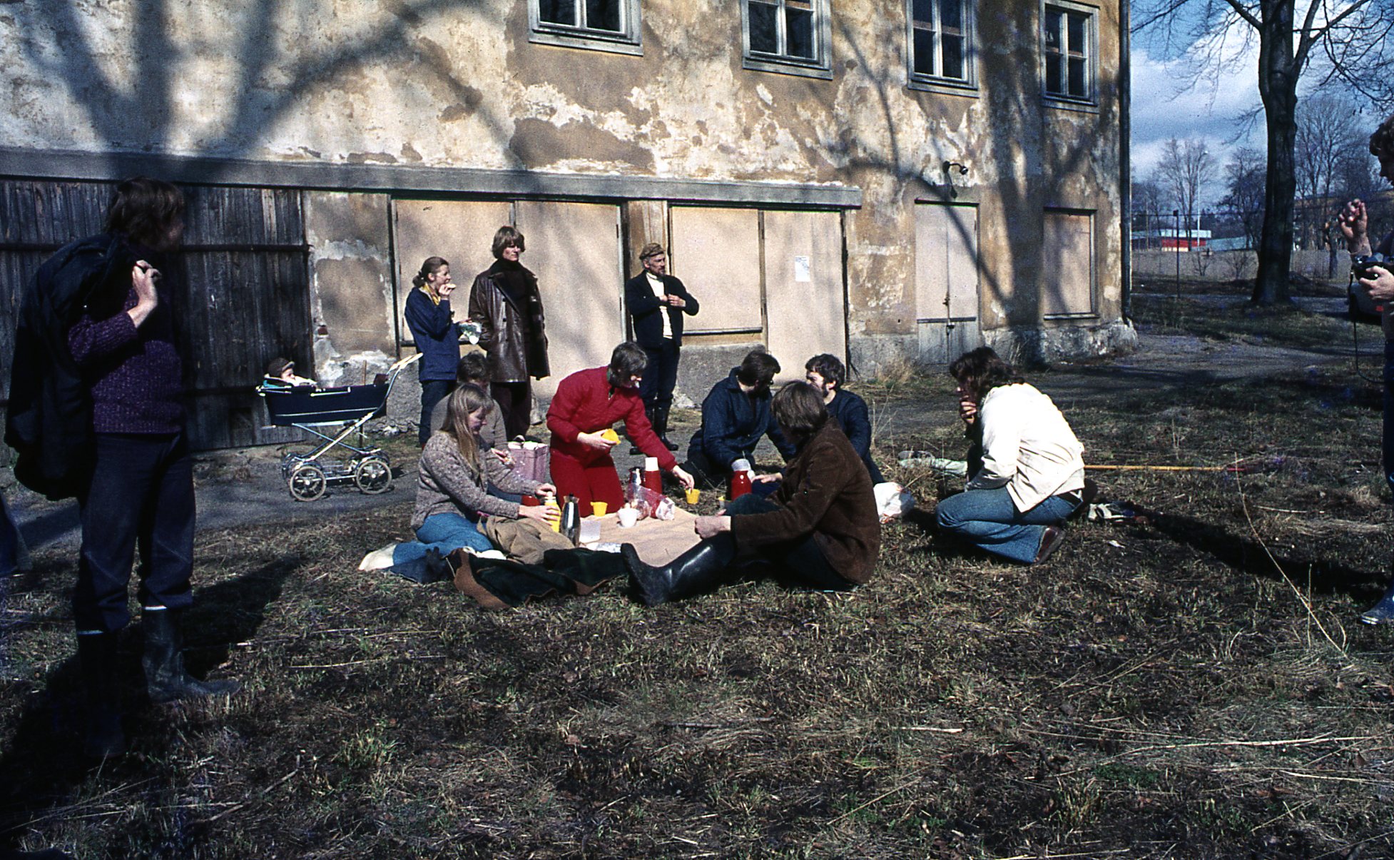 12 personer, varav en i barnvagn vid herregårdens flagnade fasad med igenspikade dörrar. Några har satt sig i gräset med kaffetermosar.
