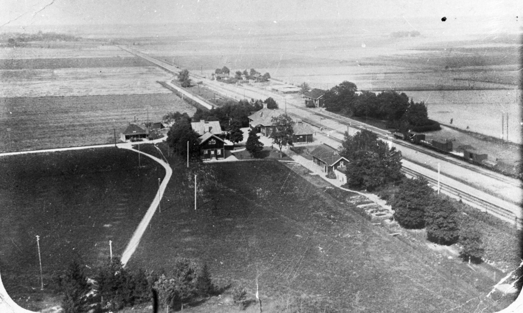 Svartvitt foto som visar järnvägen med stationshus och en handfull andra byggnader nära spåret. Resten är åker och äng.