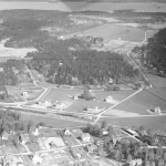 Svartvitt flygfoto med järnväg som går genom ett öppet landskap med enstaka villor.