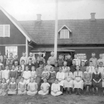 Barnen uppställda framför skolhuset med läraren i hatt i centrum.