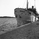 Ett svartvitt foto av en stor vit båt vid kajen. I fören står namnet skrivet.