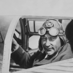 Närbild av en man som sitter i en flygplanshytt med skinnmössa och flygarglasögon.