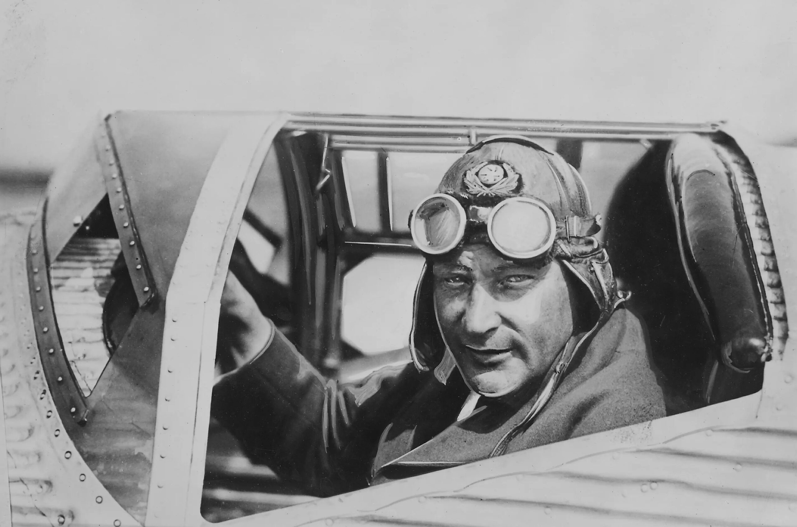 Närbild av en man som sitter i en flygplanshytt med skinnmössa och flygarglasögon.