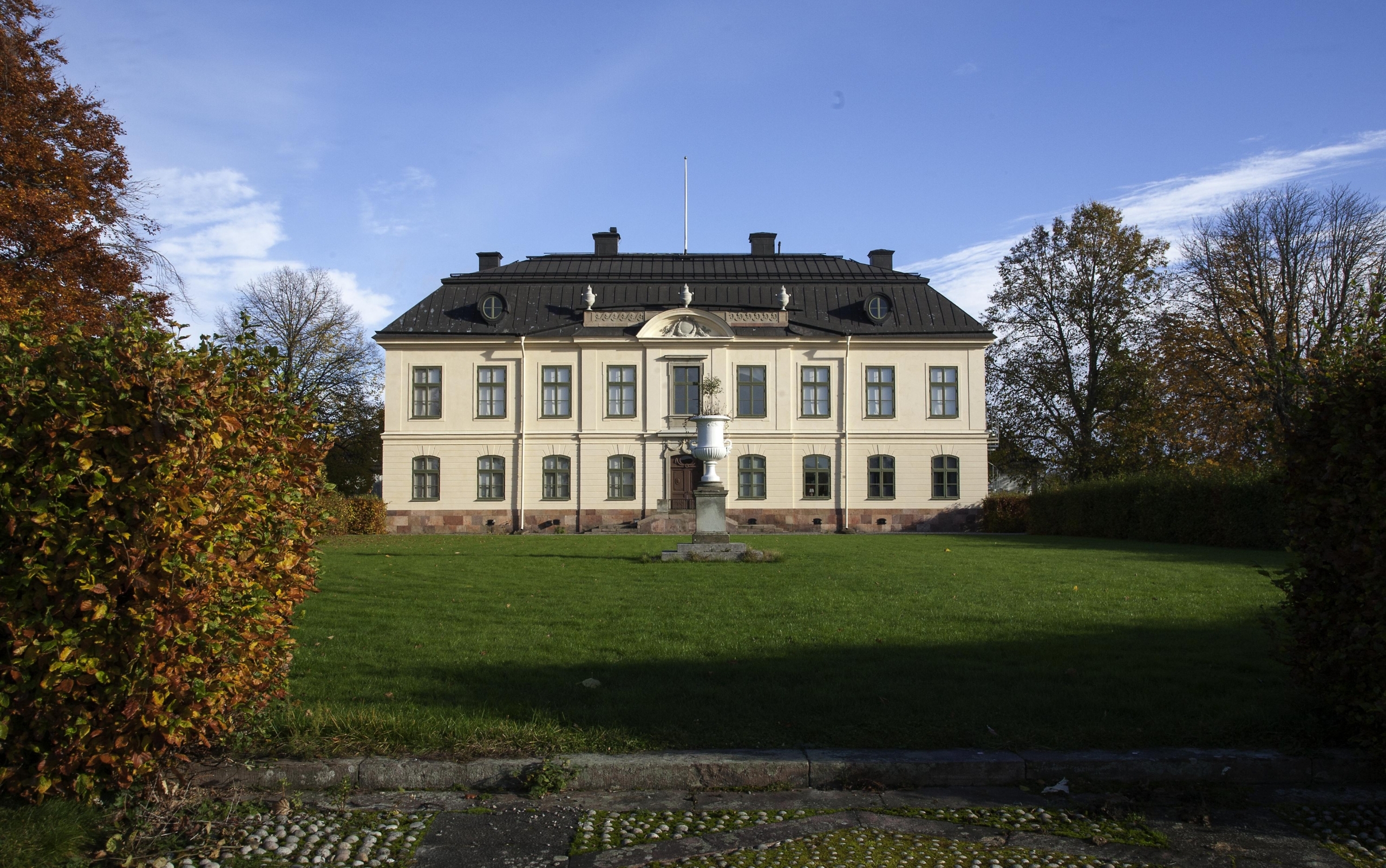 Stor, gul herrgårdsbyggnad med svart tak. Framför herrgården ligger en stor välklippt gräsmatta