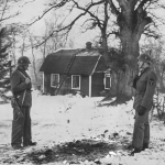 Svartvitt foto där två militärer står på var sin sida av en svart fläck i den vita snön, med stugan i bakgrunden.