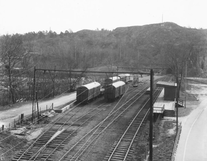 Ett svartvitt foto där godsvagnar står på ett av de fyra spåren i spårområdet. Spåren försvinner in i en tunnel i berget och inga hus syns.