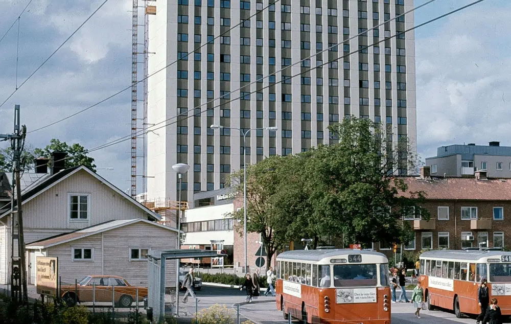 Ett äldre trähus med ett stort höghus i bakgrunden. Två äldre bussar visar att bilden är från 1900-talet.