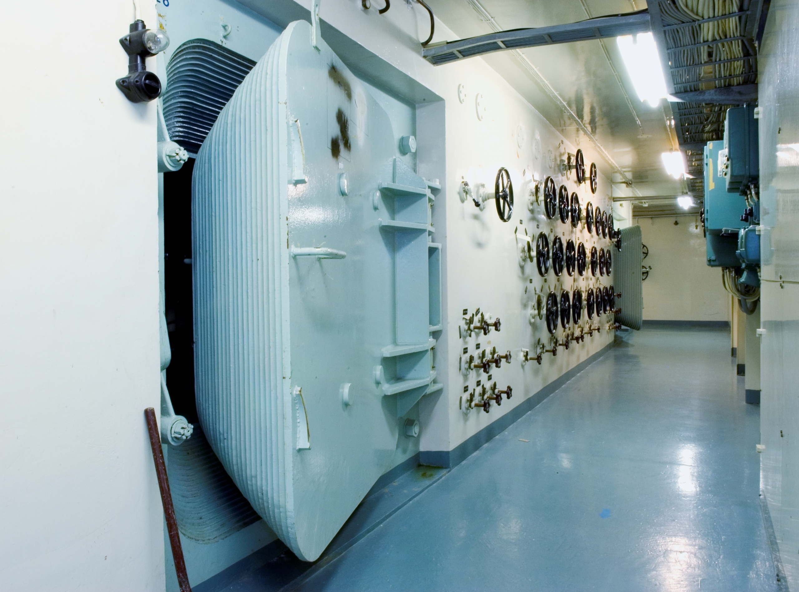 En mycket tjock dörr står på glänt i en korridor där väggen är full av rattar och kranar.