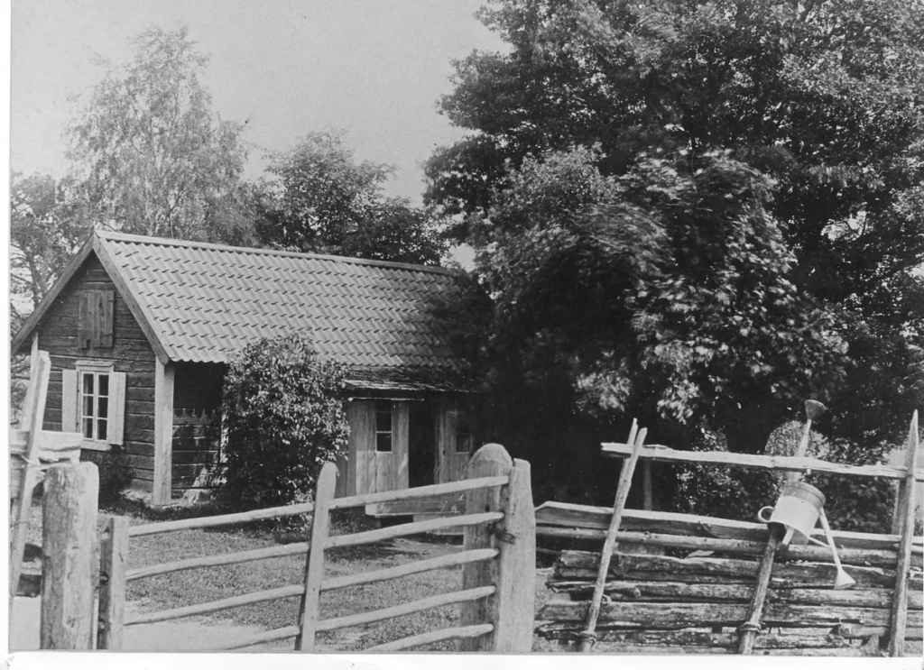 Svartvitt foto av en mindre stuga bredvid ett stort träd.