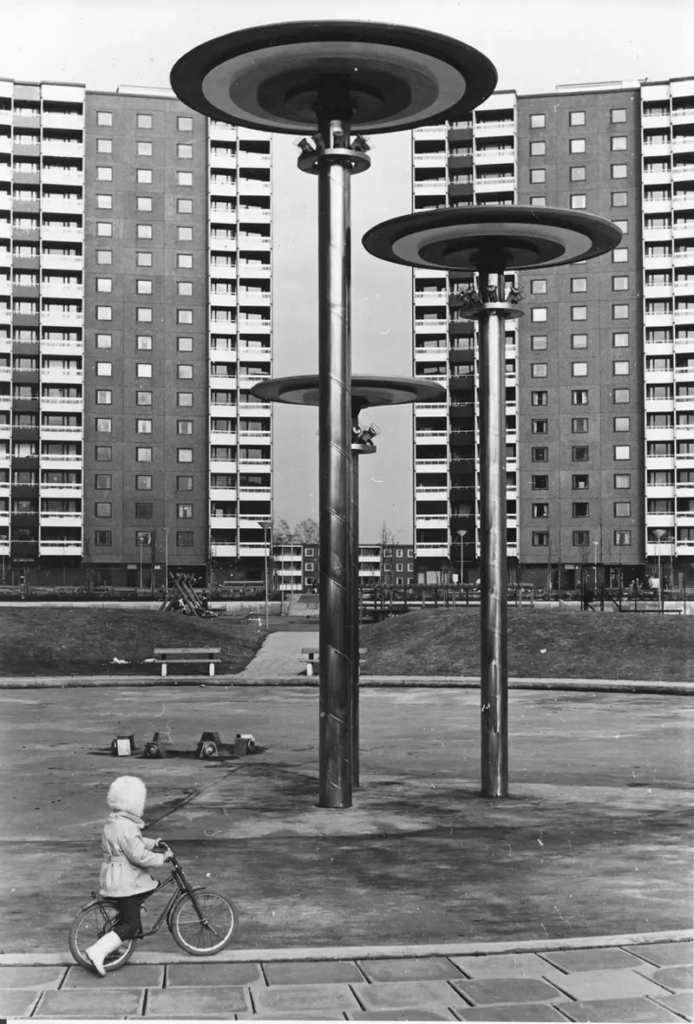 Svartvitt foto. Barn cyklar bland skulpturer utanför höghus.